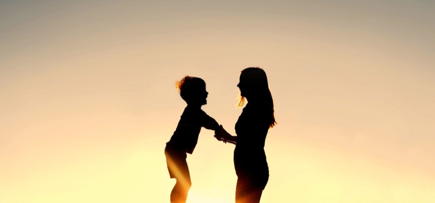 Silhouette of a young mother lovingly holding hands with her happy little child outside in front of a sunset in the sky.