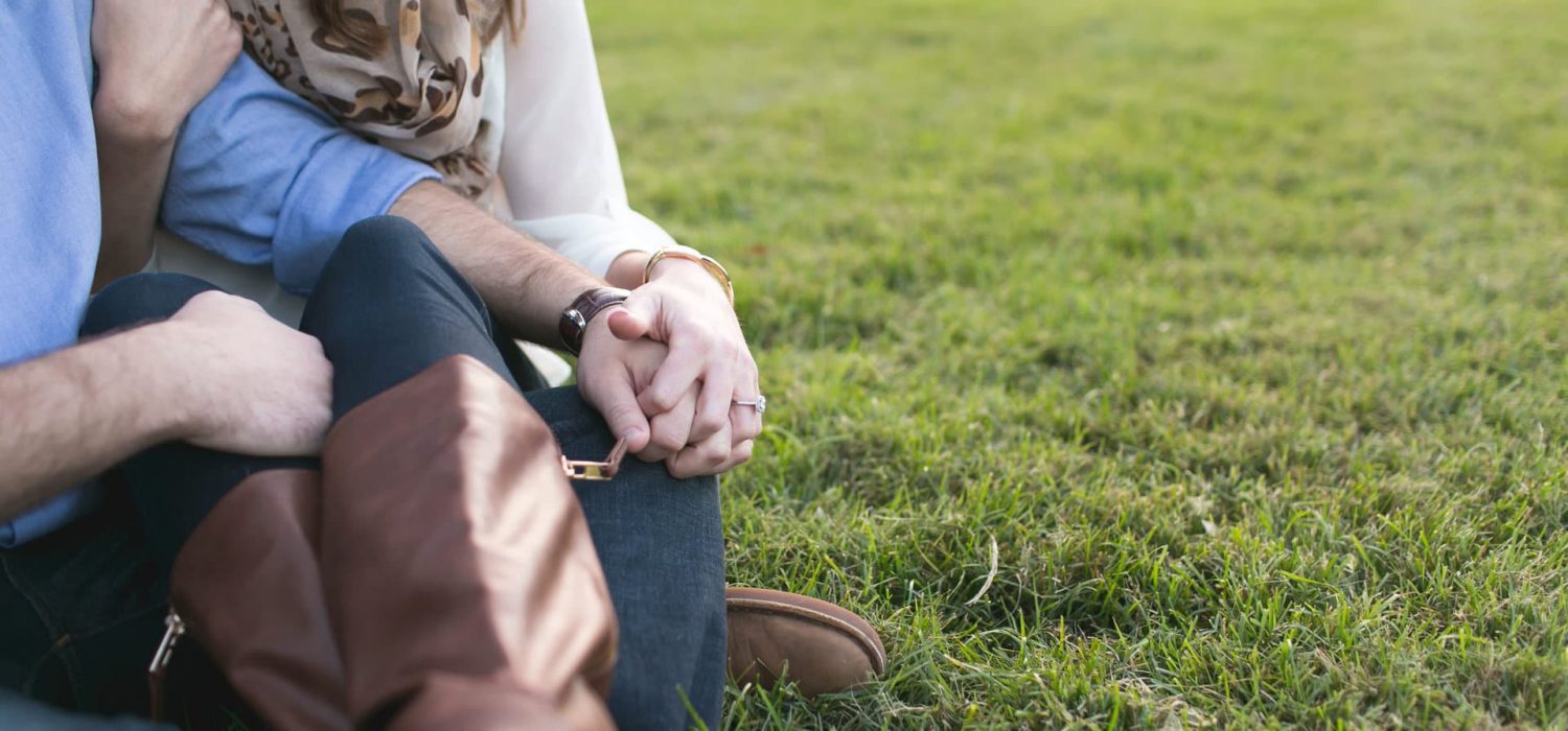 e-session-couple-sitting-in-grass-holding-hand-ring-real-moment-engagement-session-picture